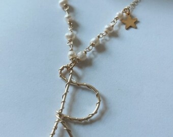 Puede incluir: Un collar de oro con un dije de perla y estrella. El collar presenta un dije de alambre en forma de letra 'R'.