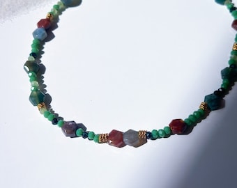 Puede incluir: Un collar de cuentas con una variedad de piedras preciosas de colores. El collar tiene un cierre y detalles de alambre de color dorado. Las piedras preciosas son en tonos verdes, rojos, azules y grises. El collar está sobre una superficie blanca.