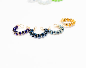 Puede incluir: Cuatro anillos envueltos en alambre con cuentas de cristal de diferentes colores. Los anillos están hechos con alambre dorado y presentan cuentas azules, moradas, verde azulado y doradas.