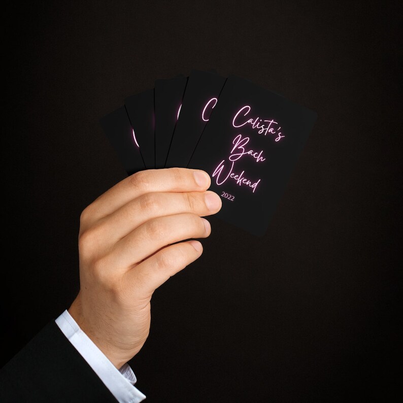 May include: A hand holds a stack of black cards with pink text that reads "Calista's Bach Weekend 2022".