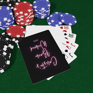 May include: A black card with pink neon text that reads "Cards & Drinks" and "Weekend 2023". The card is on top of a stack of playing cards, including a Jack of Spades, a Jack of Diamonds, a 10 of Diamonds, and a 10 of Spades. The cards are on a green felt surface with red and blue poker chips.