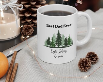 May include: White ceramic mug with a black and green design. The mug says "Best Dad Ever" and features a row of pine trees. The names Kaylan, Johnny, and Serena are written below the trees.