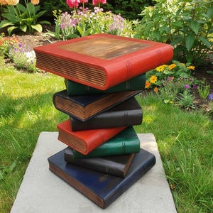 May include: A sculpture of stacked books in a garden setting. The books are painted in shades of red, green, black, and blue, and rest on a grey stone slab. The background features green grass and a variety of colorful flowers.