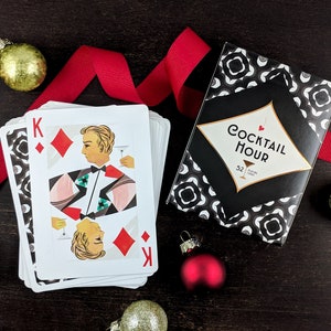 Cocktail Hour Playing Cards showing the King of diamonds on top of the stack of cards with the tuck box besides it, and surrounded by a festive red holiday ribbon and ornaments.