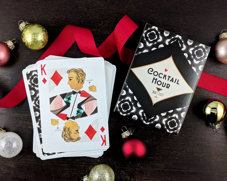 Cocktail Hour Playing Cards showing the King of diamonds on top of the stack of cards with the tuck box besides it, and surrounded by a festive red holiday ribbon and ornaments.