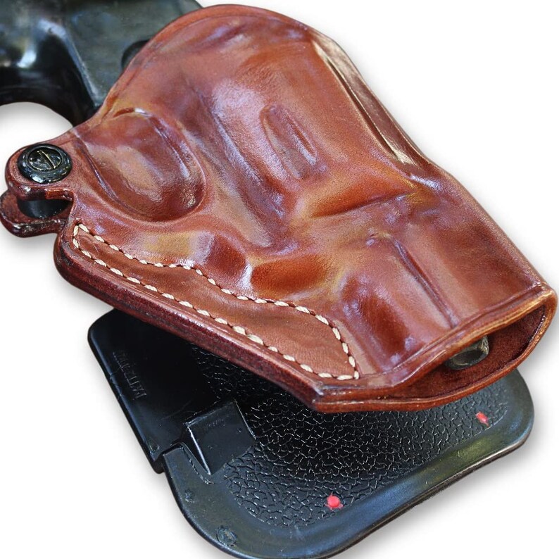 May include: A brown leather holster with white stitching for a handgun. The holster is attached to a black plastic base.