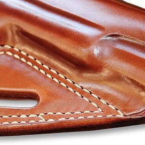 May include: A brown leather holster with white stitching. The holster has a belt loop and a cut-out for a gun.