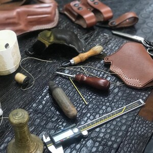 May include: A leatherworking workspace with tools and materials, including a brown leather holster, a spool of thread, a thimble, a awl, a ruler, and a piece of alligator skin.
