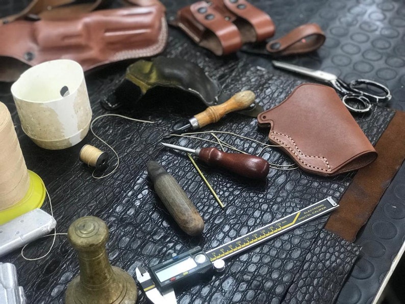 May include: A leatherworking workspace with tools and materials, including a brown leather holster, a spool of thread, a thimble, a awl, a ruler, and a piece of alligator skin.