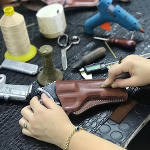 K&ouml;nnte beinhalten: Eine Person arbeitet an einem braunen Lederholster f&uuml;r eine Waffe. Das Holster wird mit Nadel und Faden gen&auml;ht. Auf dem Tisch liegen weitere Werkzeuge, darunter eine Klebepistole, eine Garnrolle und ein Messwerkzeug.