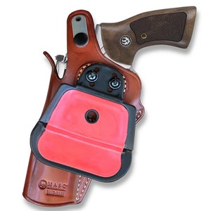 May include: A brown leather holster with a black plastic clip and a red plastic insert. The holster is designed to hold a revolver. The holster is made by MASC International Hand-Made.