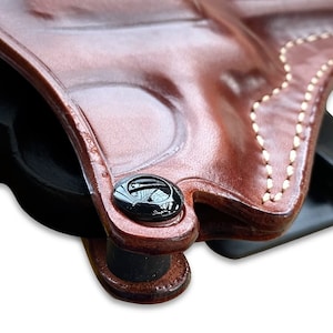 May include: Close-up of a brown leather holster with a black snap button. The holster is stitched with white thread.