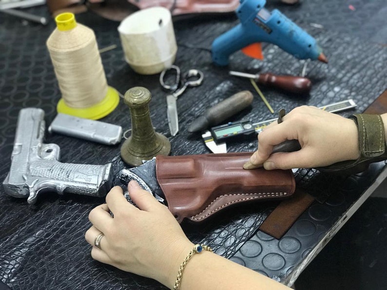 May include: A person working on a brown leather holster for a gun. The holster is being stitched with white thread. The person is using a tool to smooth out the leather.