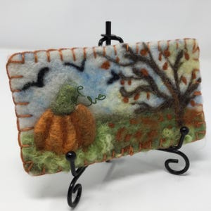 May include: A felt wall hanging with a pumpkin, a tree with brown branches and orange leaves, and a blue sky with flying birds. The felt piece is on a black easel.
