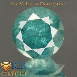 Puede incluir: Una gema redonda de color verde azulado con un patrón de estrella de facetas blancas. La gema está certificada por IGR. El texto en la parte superior dice "See Video in Description".