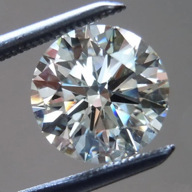 May include: A round-cut diamond held by a pair of tweezers. The diamond is clear and reflects light, showcasing its facets. The tweezers are silver-colored.