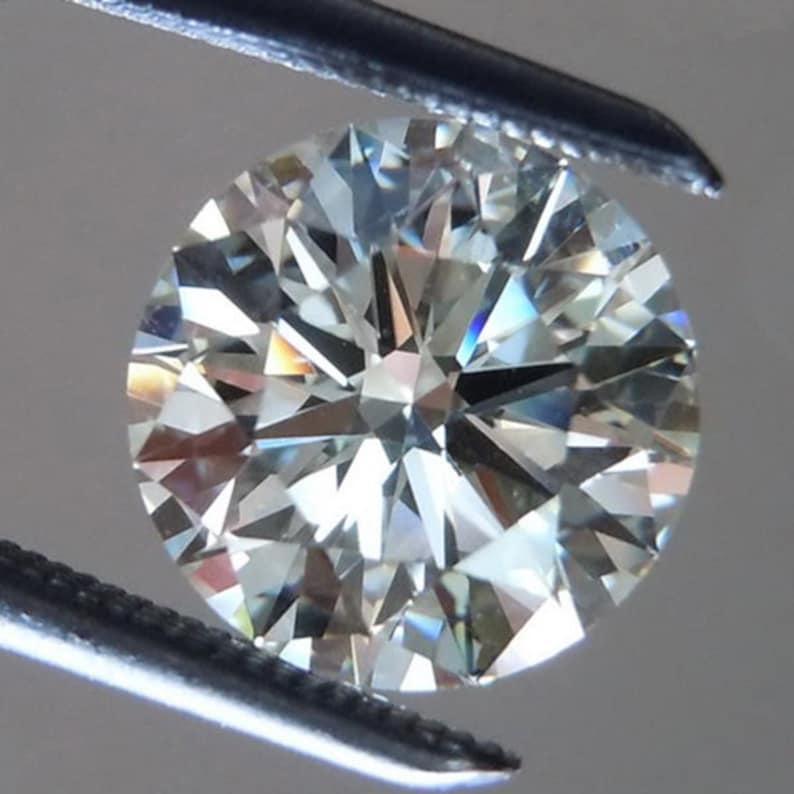 May include: A round-cut diamond held by tweezers. The diamond has many facets that reflect light, creating a sparkling effect. The gemstone appears colorless with hints of blue and yellow.
