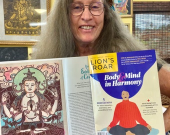 Op de afbeelding: Een vrouw houdt een open magazine vast met de titel "Lion's Roar: Body & Mind in Harmony". De omslag van het magazine toont een illustratie van een persoon die mediteert in een rood shirt. Het magazine is opengeklapt naar een artikel over "5 Meditaties voor meer ontspanning, bewustzijn en onvoorwaardelijke liefde".