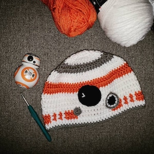 May include: A white, orange, and gray crocheted hat with a black circle design. The hat is shaped like a character from the Star Wars movie franchise.