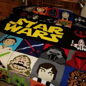 May include: A black crochet blanket with a yellow "STAR WARS" logo in the center. The blanket is made up of squares with different Star Wars characters, including Yoda, Darth Vader, and Princess Leia.