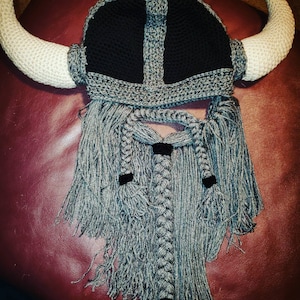 Puede incluir: Un gorro de vikingo de ganchillo con barba gris y cuernos. El gorro es gris y negro con una barba trenzada.