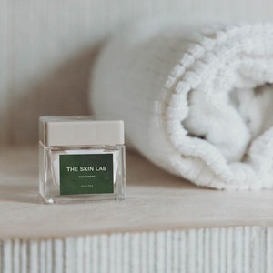 May include: A clear glass jar with a beige lid and a green label that reads "THE SKIN LAB BODY CREME". A rolled white towel is in the background. The jar contains 59 g of product.