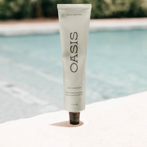 May include: A white tube of Oasis Daily Sunscreen with the text "FOR ALL SKIN TYPES" and "Mineral Body Sunscreen Broad Spectrum SPF 50" printed on the tube. The tube is 1.7 oz / 50g.