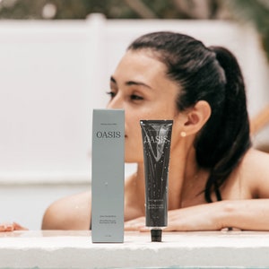 May include: Two gray tubes of Oasis Daily Sunscreen, one is a full-size bottle and the other is a travel-size tube. The tubes are sitting on a white surface near a pool.