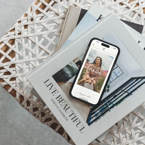 May include: A smartphone displaying a photo of a woman in a pink dress, with the text "STUDIO FULCHI" on the screen, rests on a stack of books. The books are titled "LIVE BEAUTIFUL" and "K". The items are on a woven chair.