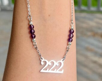 May include: A silver-toned necklace featuring a central pendant shaped as the number 222. The chain includes two sections of three small, round, deep purple amethyst beads symmetrically placed on each side of the central numeric pendant.