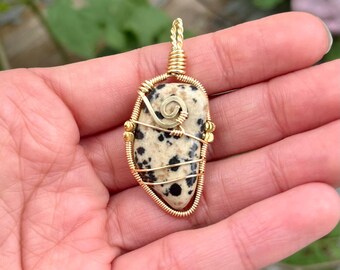 May include: A pendant featuring a smooth, cream-colored stone with black spots, encased in a gold-toned wire wrap. The wire forms a decorative spiral detail on the front and extends into a twisted loop at the top for hanging the jewelry piece.