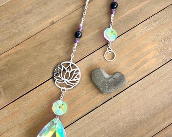 May include: A decorative suncatcher with a silver lotus flower charm, iridescent crystal teardrop, and assorted beads in purple, teal, and black. A small gray heart-shaped stone is also present. The suncatcher hangs from a silver chain.
