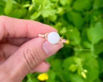 May include: A wire-wrapped ring featuring a single, smooth, round white stone set in a copper-colored metal band. The band is constructed from thin, coiled wire that secures the central stone in place with a simple, symmetrical design.