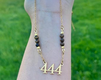 May include: A gold-toned necklace featuring a pendant with the number 444 in a serif font. The chain is accented with two sections of three dark brown, spherical beads separated by small gold-toned spacers, creating a balanced, layered look.