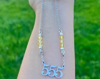 May include: A silver-toned necklace featuring a pendant with the number 555 in a stylized font. The chain includes two sections of three small, translucent yellow beads separated by silver-toned spacers, positioned symmetrically on each side.