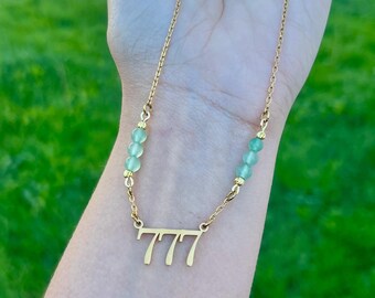 May include: A gold-toned necklace featuring a central pendant with the number 777 in a stylized font. The chain is accented on both sides with three small, round, translucent light green beads separated by tiny gold-toned metal spacers.