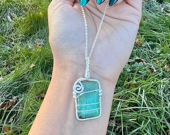 May include: A rectangular green stone pendant encased in silver-colored wire wrapping. The pendant features a spiral wire detail at the top left corner and is suspended from a thin silver-colored chain necklace.