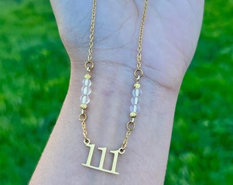 May include: A gold-toned necklace featuring a pendant with the number 111 in a serif font. The chain includes two sections of three small, clear, spherical beads separated by tiny gold-toned spacers, connected to a delicate gold-toned link chain.