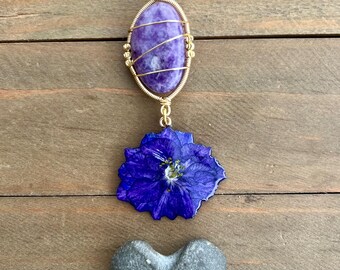 May include: A gold-colored chain necklace with a purple oval stone pendant wrapped in gold wire. Below the pendant is a blue flower and a gray heart-shaped stone. The necklace is displayed on a wooden surface.