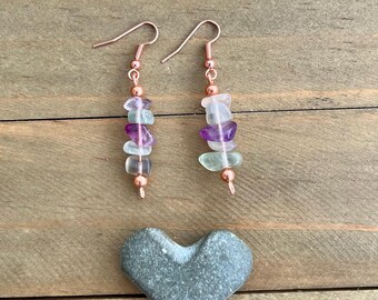 May include: A pair of handcrafted earrings with a copper-colored hook and small beads. The earrings feature a vertical arrangement of colorful, irregularly shaped stones in shades of purple, green, and clear. A gray heart-shaped stone rests below.