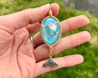 May include: A handcrafted pendant featuring a blue, oval-shaped glass stone with a wavy design, encased in gold-colored wire. A small, dark gray and gold-colored whale tail charm hangs below. The pendant is held in a hand against a blurred green background.