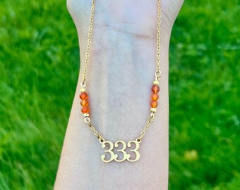 May include: A gold-toned necklace featuring a central pendant with the number 333 in a stylized font. The chain is accented on both sides by three small, round, translucent orange beads, creating a symmetrical design with a delicate link chain.