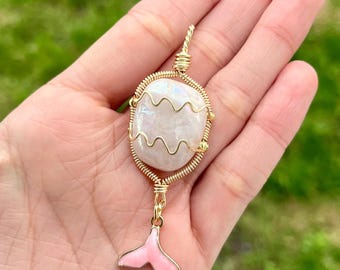 May include: A handmade pendant featuring a pearlescent, wavy-edged stone encased in gold wire. A pink enamel mermaid tail charm dangles below. The pendant is held in a hand against a blurred green background.