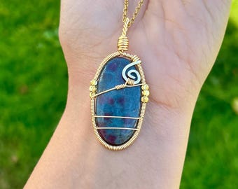 May include: An oval-shaped pendant featuring a dark blue and red speckled stone encased in gold-toned wire wrapping with a silver-toned spiral accent, suspended from a matching gold-toned chain necklace.