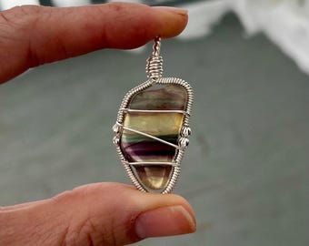 May include: A handcrafted pendant featuring a polished, multi-colored fluorite stone wrapped in silver-colored wire. The stone displays bands of purple, green, and yellow hues. The pendant is held between two fingers against a blurred background.