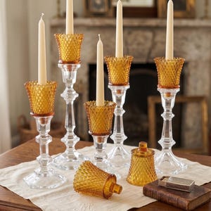 K&ouml;nnte beinhalten: Eine Sammlung von Kerzenhaltern aus klarem Glas mit bernsteinfarbenen Glaseins&auml;tzen im Rautenmuster. Vier Halter enthalten lange, d&uuml;nne, cremefarbene Stabkerzen. Zwei weitere bernsteinfarbene Glaseins&auml;tze liegen neben einer kleinen, rechteckigen Schachtel.