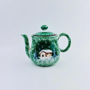 May include: Green ceramic teapot with a winter scene featuring a snow-covered house and trees. The teapot has a lid with a small knob and a curved handle. The design is painted on the body of the teapot.
