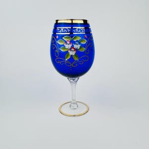 May include: A cobalt blue wine glass with a gold rim and intricate floral designs painted on the side. The glass has a clear stem and a gold-rimmed base.