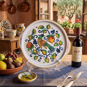 May include: A round ceramic serving platter with handles, featuring a hand-painted design of a central bird surrounded by colorful fruit and swirling blue vines. The platter has a white background with a yellow rim and is displayed on a clear stand.
