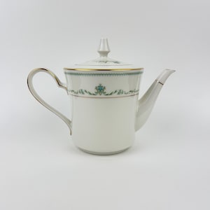 Może przedstawiać: Biały porcelanowy imbryk z pokrywką, uchwytem i dzióbkiem. Imbryk ma złoty brzeg i ozdobny pas z turkusowymi wzorami kwiatowymi. Uchwyt i dzióbek mają złote akcenty.